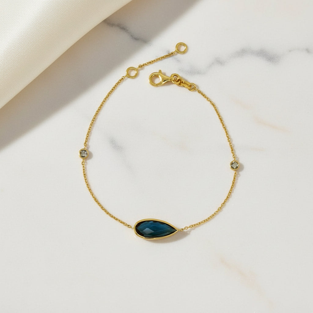 Feines Goldarmband mit einem dunkelblauen, tropfenförmig facettierten Mittelstein und zwei kleinen runden Akzentsteinen in zarter Kette, mit Karabinerverschluss und Verlängerungskettchen.