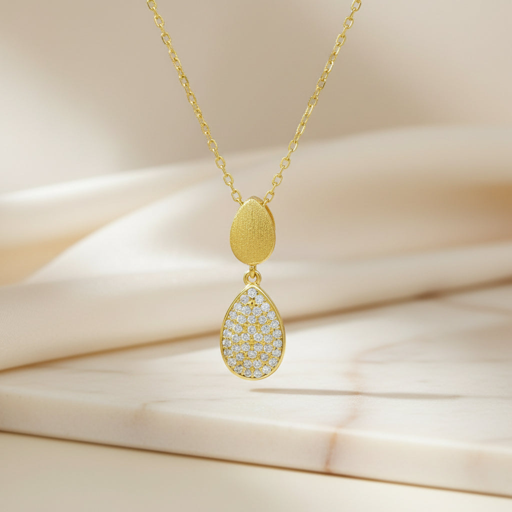 Detailansicht eines vergoldeten Doppel-Tropfen-Anhängers, oben matt strukturiert und unten vollflächig mit klaren Zirkonia-Steinen ausgefasst.