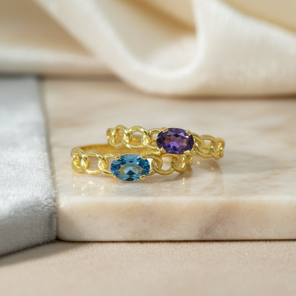 Zwei Gelbgoldringe R34 nebeneinander, einer mit blauem London Topas, der andere mit violettem Amethyst, beide mit Kettenglieder-Optik.
