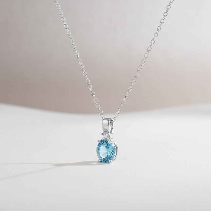 Feines Silbercollier mit kleinem rund facettiertem blauem Topas-Anhänger auf zarter Ankerkette vor weißem Hintergrund.